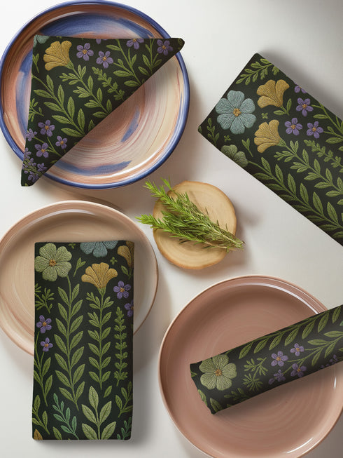 A 4-piece cloth napkin set featuring embroidered-style botanical illustrations of wildflowers and herbs in shades of gold, mint, and lavender on a dark green background; made from soft polyester, square-shaped, with a luxe and mystical design.